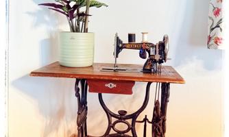 An old sewing machine stands on a wooden floor, surrounded by a potted plant in a light flower pot. The name "PAULA" is visible in the image.
