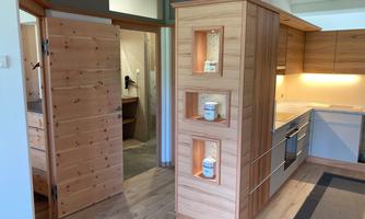 A modern kitchen with wooden paneling and an open floor plan. In the background, a door to a bathroom is visible.