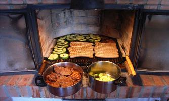 A grill with various types of meat and vegetables on the grate. Two pots with cooked meat and another side dish are standing next to it.
