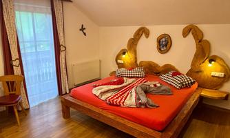 A cozy bedroom with a large wooden bed and colorful blankets. The room has a large window and rustic decorative elements.