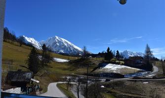 A picturesque landscape with snow-covered mountains and a clear blue sky. The path winds through green meadows and forests.