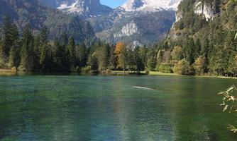 A clear lake surrounded by tall mountains and dense forests. The sky is blue with some clouds.