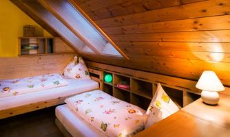 A cozy attic room with two single beds and a wooden ceiling. There is a bedside lamp and an open shelving unit.