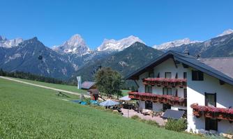 A picturesque building with balcony flowers in a green meadow. In the background, impressive mountains and a clear blue sky can be seen.