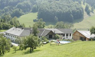 A rural landscape with several buildings and a swimming pool. In the background, wooded hills and meadows can be seen.