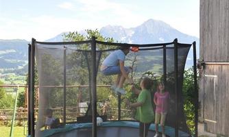A trampoline stands outdoors, surrounded by green meadows and mountains in the background. Children are happily jumping on it and enjoying the day.