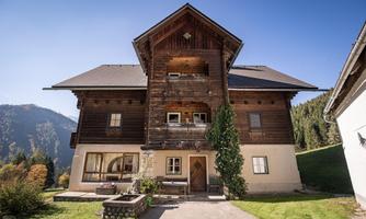 A large, rustic wooden house in the mountains. The surroundings are green and there is a clear blue sky.
