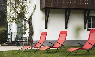 Red loungers are on green grass next to a white house. In the background, a tree and a porch are visible.