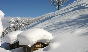 A winter landscape with snow-covered hills and a blue sky. Two snow-covered cabins lie in the foreground.