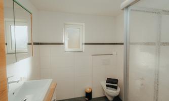 A modern bathroom with a shower, sink, and toilet. Large windows provide plenty of natural light.
