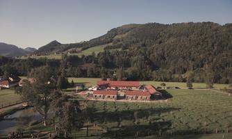 A picturesque landscape with gentle hills and lush greenery. In the foreground stands a building with red roofs, surrounded by meadows and a small pond.