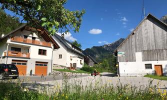 A quiet rural road with two houses and green meadows. In the background, mountains and a blue sky are visible.