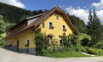 A yellow house with a red roof and many windows. Surrounding the house are green trees and flowers.