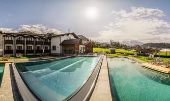 A modern hotel with a large outdoor pool and sun loungers. The landscape is green and the mountains are visible in the background.