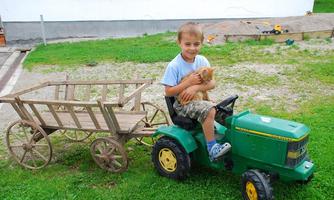 A boy is sitting on a small green tractor and holding a kitten in his arms. Next to him is a wooden cart on the meadow.