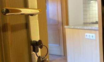 A door with a key and a lock that leads to a room. In the background, a bright kitchenette is visible.