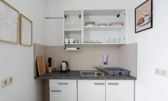 A small, modern kitchen with white cabinets and a gray countertop. Above, glasses and dishes are neatly arranged, and a picture hangs on the wall.