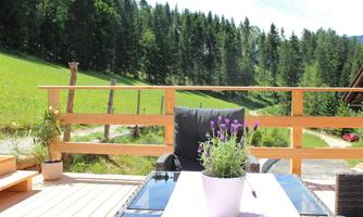 A cozy terrace with a table and a pot full of lavender flowers. In the background, green meadows and dense forests are visible.