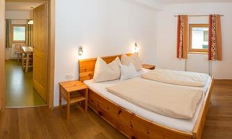 A cozy bedroom with a double bed and wooden dressers. In the background, a table and chairs are visible, suggesting a dining area.
