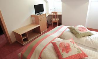 A cozy bedroom with a double bed and colorful cushions. In the background, there is a desk with a television.