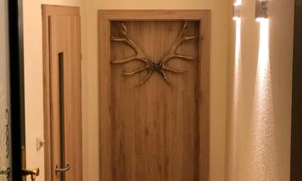 A narrow corridor with light walls and a wooden door featuring a antler decoration. On the right, two wall sconces are mounted, illuminating the space.