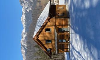 A cozy wooden house in the snow with mountains in the background. The sky is clear and blue.