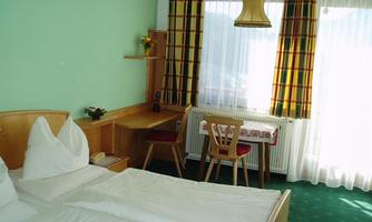 A bright room with two beds and a wooden desk. Large windows provide plenty of daylight and create a cozy atmosphere.