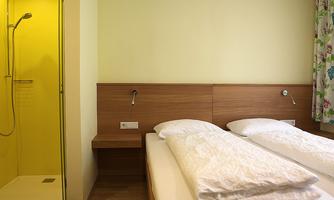 A modern room with two single beds and bright walls. On the left side, there is a shower cubicle with yellow walls.