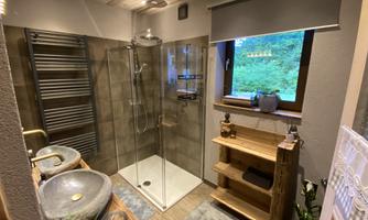 A modern bathroom with a glass shower and two stone sinks. Large windows provide plenty of daylight and a view of the greenery.
