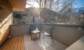 A sunny balcony with two chairs and a small table. In the background, mountains and trees can be seen.