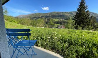 A cozy terrace with two blue chairs and a beautiful view of green meadows and mountains. The sky is clear and blue.