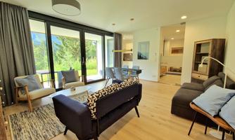 A modern furnished living room with large windows and a view of the greenery. There are comfortable furniture and a bright, inviting space.
