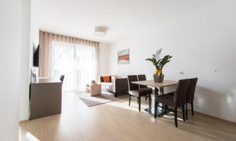 A modern living room with light wood flooring and large windows. There is a cozy seating area, a dining table, and a potted plant.