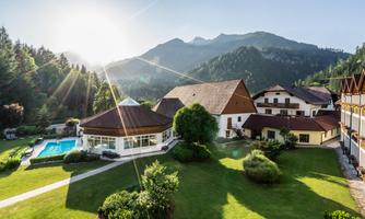 A picturesque view of a resort in the mountains, surrounded by green landscape. In the foreground, there is a pool and the sun is shining brightly.