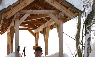 A man is sitting with a dog in the snowy landscape under a wooden shelter. The surroundings are wintry and covered in snow, with trees in the background.