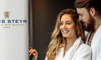A couple in bathrobes smiles relaxed and enjoys the moment. In the background, the logo of the Vitus Steyr Hotel can be seen.