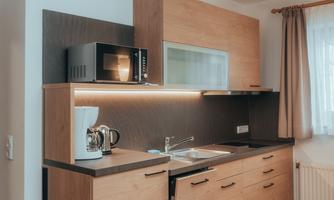 A modern kitchen with wooden shelves and a built-in microwave. It features a sink, coffee machine, and large windows for plenty of light.