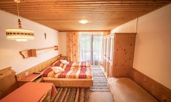A cozy hotel room with wooden floors and warm colors. There is a double bed, a table, and a wardrobe, as well as large windows with natural light.