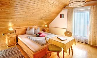 A cozy bedroom with wooden paneling and a large window. There is a double bed, a table, and two chairs.