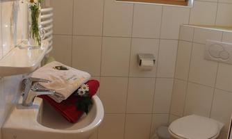 A small bathroom with white tiles and a sink. On the floor is a red bath towel, and there is a window that lets in daylight.