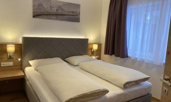 A cozy bedroom with a double bed and two nightstands. The room has light walls, a window with curtains, and a decorative picture on the wall.