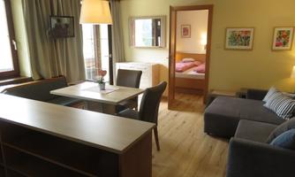 A cozy apartment with a living area, table, and chairs. In the background, there is a bedroom with a double bed and colorful pictures on the walls.
