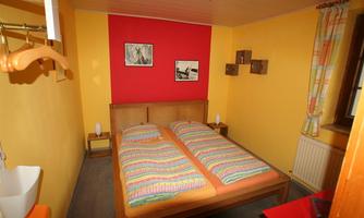 A cozy bedroom with a double bed and colorful bedspreads. The walls are painted in vibrant colors, and there are decorative elements like pictures and shelves.