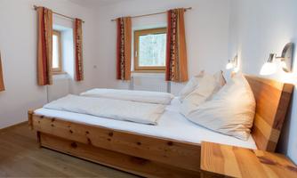 A cozy bedroom with a wooden bed and two pillows. The walls are light and there are large windows with curtains.
