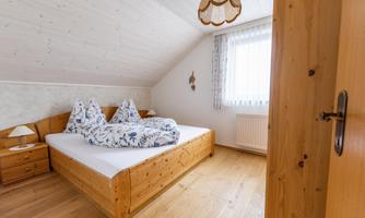 A bright bedroom with a double bed and wooden dressers. Large windows provide pleasant light.