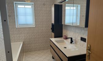A modern bathroom with a bathtub, a simple sink, and plenty of mirror surface. The walls are tiled, and a window provides daylight.