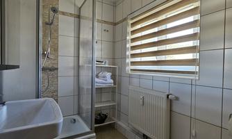 A modern bathroom with a shower cabin and a sink. Windows with blinds provide daylight.