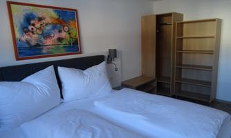 A modern bedroom with a double bed and large, white pillows. A colorful picture hangs on the wall, and there is a wardrobe and a shelf in the room.