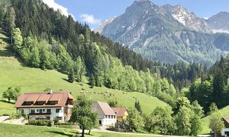 An idyllic landscape with gentle hills and green trees. In the background, majestic mountains rise under a blue sky.