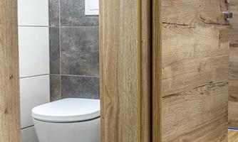 A modern bathroom area with a white toilet and a wooden door frame. The floor is made of gray tiles and wood.